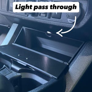 May include: A black car interior with a compartment labeled "Light pass through." The compartment is open, revealing a small storage space. An arrow points to the opening. The dashboard has buttons and a power outlet.