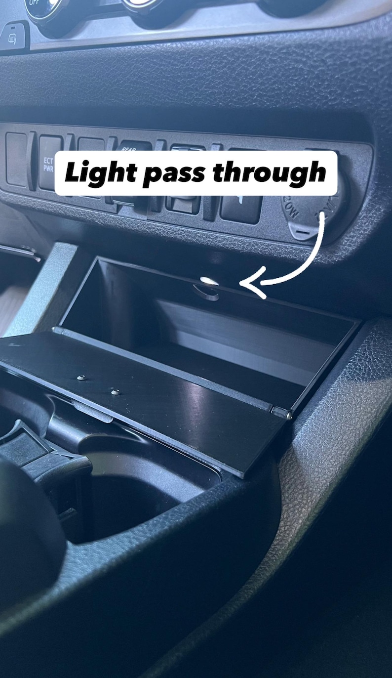 May include: A black car interior with a compartment labeled "Light pass through." The compartment is open, revealing a small storage space. An arrow points to the opening. The dashboard has buttons and a power outlet.