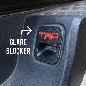 May include: A black plastic glare blocker with a red "TRD" logo. The glare blocker is attached to the back of a vehicle.