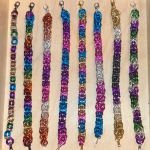 Könnte beinhalten: Neun bunte Armbänder aus Metallkettengliedern. Die Armbänder sind in verschiedenen Farben, darunter Lila, Blau, Grün, Gold und Silber.