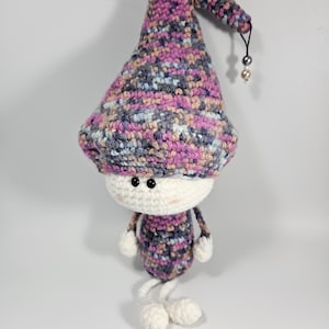 May include: A crocheted gnome doll with a pink, gray, and blue patterned hat and body. The gnome has white yarn for arms and legs and black button eyes.