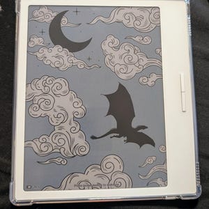 Peut inclure: Une liseuse électronique avec un cadre blanc et un étui de protection transparent. L'écran affiche un fond gris avec un motif de dragon noir, de lune et de nuages. Le texte "Press power button to wake up" est visible.