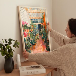 Puede incluir: Una impresi&oacute;n art&iacute;stica enmarcada que representa el interior de un invernadero soleado lleno de plantas y flores en macetas. La obra de arte presenta una paleta de colores vibrantes de rosas, naranjas y verdes, con un marco blanco. Una persona sostiene la impresi&oacute;n.
