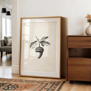 Puede incluir: Una impresión botánica en blanco y negro enmarcada de una bellota con hojas. La obra de arte se exhibe en una habitación con piso de madera y una alfombra estampada. El marco es de color madera clara.