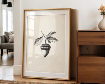 May include: A framed black and white botanical print of an acorn with leaves. The artwork is displayed in a room with a wooden floor and a patterned rug. The frame is a light wood color.
