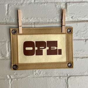 Puede incluir: Un letrero rectangular con la palabra "OPEN" en letras marrones. El letrero es beige con un borde marrón y ojales metálicos en cada esquina. Está suspendido por una cuerda y sujeto con dos pinzas de madera contra una pared de ladrillos blancos.