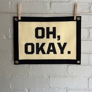 May include: A black framed banner with a cream background and the text "OH, OKAY." in black block letters.