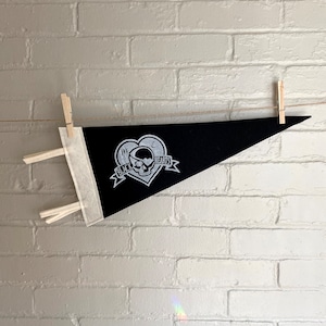 Könnte beinhalten: Schwarze Wimpelflagge mit einem weißen Totenkopf- und Knochen-Design in einem Herzen mit dem Text "Black Hearts" auf einem weißen Banner.
