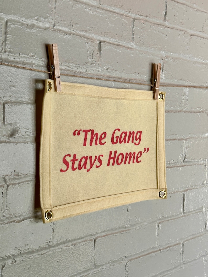 A cream felt banner with a phrase in bright pink ink reading "The Gang Stays Home" in the style of It's Always Sunny in Philadelphia.