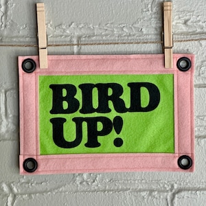 May include: A rectangular felt banner with the words "BIRD UP!" in bold black letters on a bright green background. The banner is framed in pink and has black grommets in each corner. It is suspended by wooden clothespins on a string.