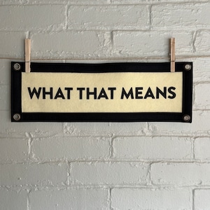 May include: A black and white banner with the text "WHAT THAT MEANS" printed in black on a beige background. The banner is hanging from a white brick wall with two wooden clothespins.