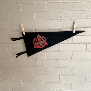 Könnte beinhalten: Schwarzer Filz-Wimpel mit einem roten gestickten Design eines Auges in einem Dreieck mit Flammen und Schlangen. Der Wimpel hängt an einer Schnur mit Holz-Wäscheklammern.