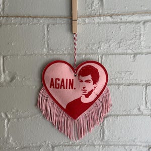 Puede incluir: Una decoración en forma de corazón rosa con la palabra «AGAIN.» y un retrato rojo de un hombre. El corazón tiene una franja en la parte inferior y está suspendido por una pinza y una cuerda contra una pared de ladrillos gris.