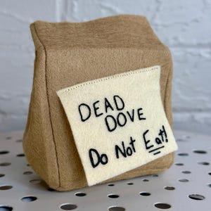 May include: A tan felt bag with a cream-colored label. The label reads "DEAD DOVE Do Not Eat!" in black lettering. The bag is sitting on a white surface with circular cutouts.