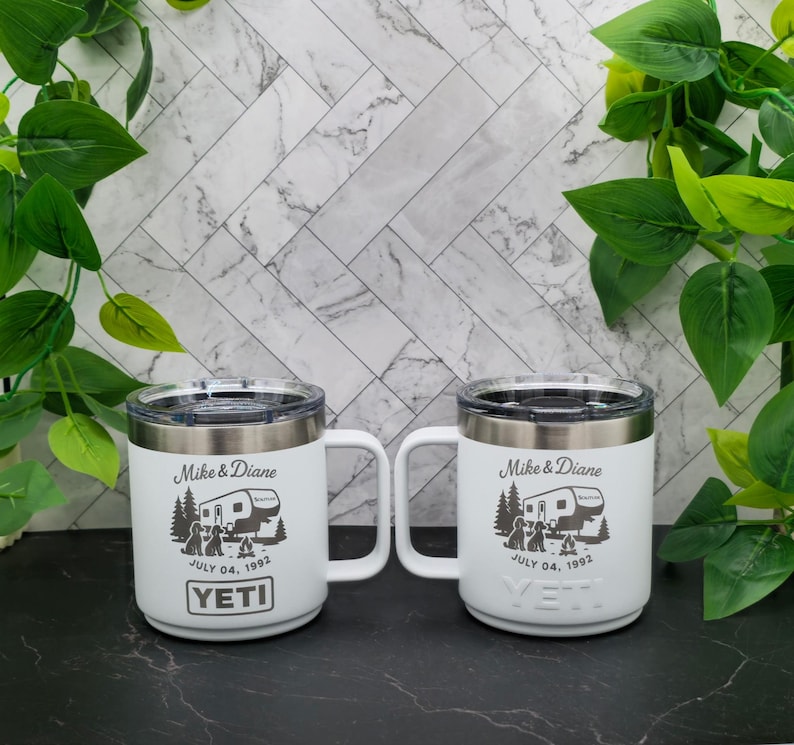Two white Yeti mugs with black handles. The mugs have a design of a camper, trees, and two dogs. The text on the mugs reads "Mike & Diane" and "July 04, 1992".
