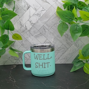 A turquoise Yeti travel mug with a stainless steel lid and handle. The mug has the text "WELL, SHIT." printed on the side.