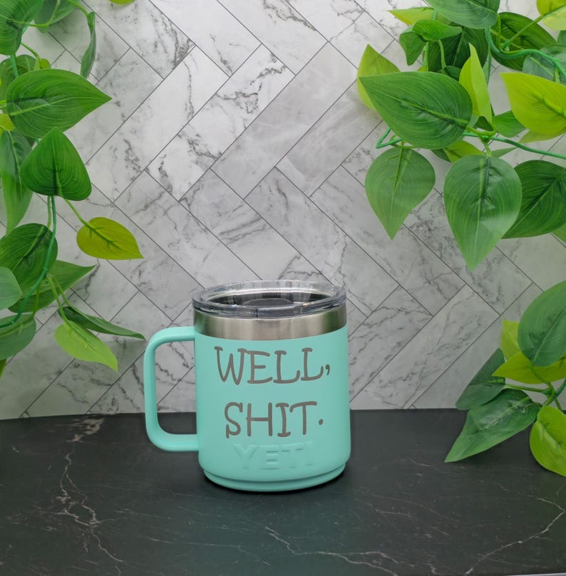 A turquoise Yeti travel mug with a stainless steel lid and handle. The mug has the text "WELL, SHIT." printed on the side.