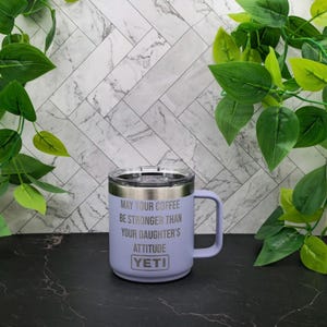 A purple Yeti travel mug with a white lid and handle. The mug has the text "May your coffee be stronger than your daughter's attitude" engraved on the side.