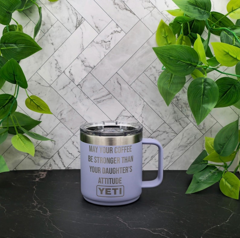A purple Yeti travel mug with a white lid and handle. The mug has the text "May your coffee be stronger than your daughter's attitude" engraved on the side.