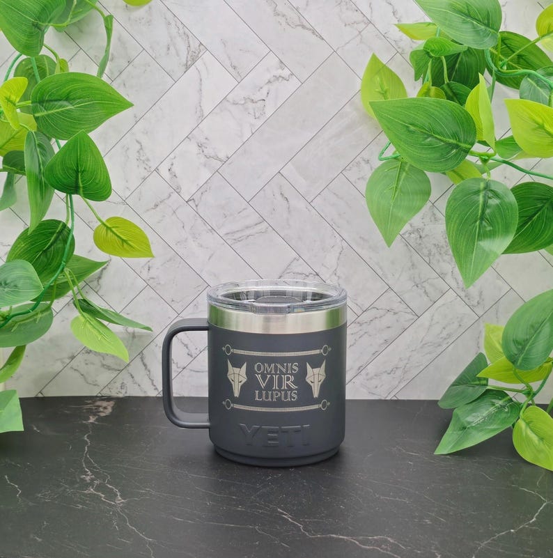 A black Yeti Rambler travel mug with a stainless steel lid. The mug has a wolf head design and the text "OMNIS VIR LUPUS" engraved on the side.
