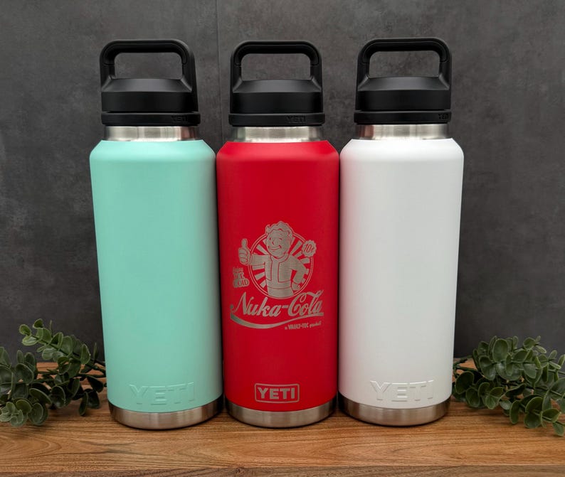 Three stainless steel water bottles with handles. The bottles are in mint green, red, and white. The red bottle has a graphic of a cartoon character on it. All bottles have the word "YETI" on the bottom.