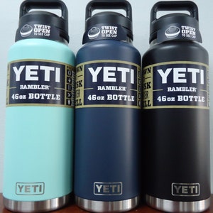 Three YETI Rambler 46oz bottles in different colors: light blue, dark blue, and black. Each bottle has a black lid with the words "TWIST OPEN TO SEE CAP" printed on it. The bottles are all stainless steel and have the YETI logo on the bottom.