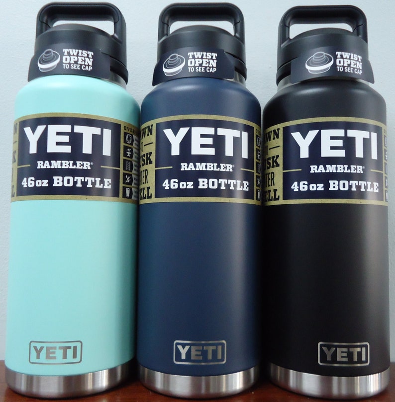 Three YETI Rambler 46oz bottles in different colors: light blue, dark blue, and black. Each bottle has a black lid with the words "TWIST OPEN TO SEE CAP" printed on it. The bottles are all stainless steel and have the YETI logo on the bottom.
