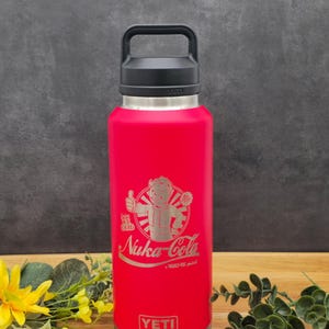 May include: Red YETI Rambler 36 oz. bottle with a black handle and lid. The bottle has a laser-etched design of a cartoon character with the text "Stay  Ice Cold" and "Nuka-Cola - A Vault-Tec product".