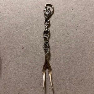 May include: A silver-toned fork with an ornate design and a grape cluster at the top. The fork has a decorative handle with a floral pattern.