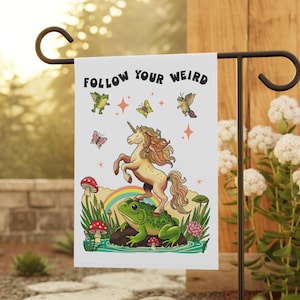 Puede incluir: Una bandera de jardín blanca con un diseño caprichoso que presenta un unicornio de pie sobre una rana, un arcoíris, setas y el texto "FOLLOW YOUR WEIRD". La bandera se muestra en un soporte de metal negro en un jardín.