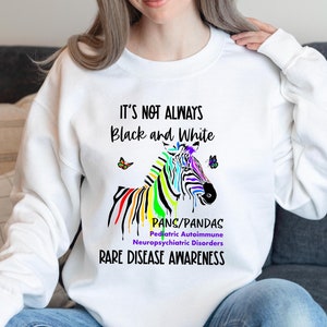 May include: White sweatshirt with the text "IT'S NOT ALWAYS Black and White" and a colorful zebra graphic. The graphic includes the text "PANS/PANDAS Pediatric Autoimmune Neuropsychiatric Disorders RARE DISEASE AWARENESS".