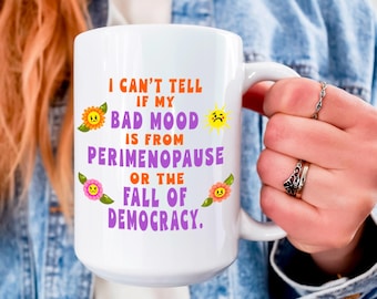 May include: A white ceramic mug featuring the orange and purple text I CAN'T TELL IF MY BAD MOOD IS FROM PERIMENOPAUSE OR THE FALL OF DEMOCRACY. The design includes small, colourful flower illustrations and a yellow sun icon with sad faces.