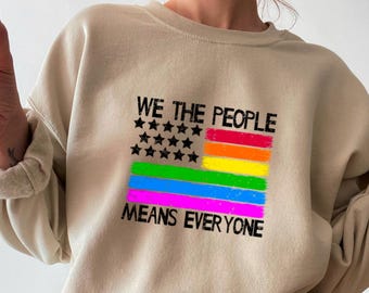 Può includere: Una felpa girocollo color beige con una stampa grafica di una bandiera stilizzata con stelle nere e strisce orizzontali rosse, arancioni, gialle, verdi, blu e viola. Il testo WE THE PEOPLE e MEANS EVERYONE è stampato in nero.