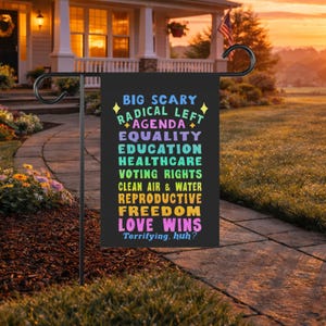 May include: A black rectangular garden flag featuring colorful text that reads: BIG SCARY RADICAL LEFT AGENDA, EQUALITY, EDUCATION, HEALTHCARE, VOTING RIGHTS, CLEAN AIR & WATER, REPRODUCTIVE FREEDOM, LOVE WINS, Terrifying, huh? The flag hangs on a black metal stand.