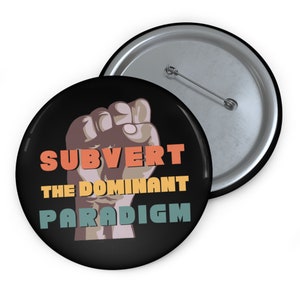 May include: A black button featuring a raised fist and the text "Subvert the Dominant Paradigm" in a retro colour scheme.
