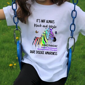 May include: White t-shirt with a colorful zebra graphic and the text "It's not always black and white. PANS/PANDAS Pediatric Autoimmune Neuropsychiatric Disorders Rare Disease Awareness."
