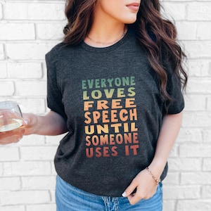 Può includere: T-shirt grigio scuro con la scritta "EVERYONE LOVES FREE SPEECH UNTIL SOMEONE USES IT" in verde, giallo, arancione e rosso. La maglietta ha un girocollo e maniche corte.