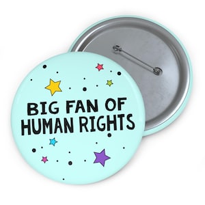 May include: A light blue button with the text "BIG FAN OF HUMAN RIGHTS" in black. The button is decorated with colorful stars and black dots. The back of the button is silver with a pin.