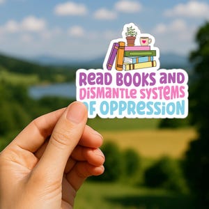 Puede incluir: Una pegatina con el texto "Read Books and Dismantle Systems of Oppression" en morado, rosa y azul. La pegatina presenta una pila de libros, una planta en maceta y una taza con un corazón. El fondo es una escena borrosa al aire libre.