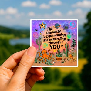 Puede incluir: Una pegatina cuadrada con el texto "The universe is experiencing and expanding through YOU." El diseño incluye una estrella, un planeta, un sol, un pulpo, cactus y un pájaro, sobre un fondo multicolor.