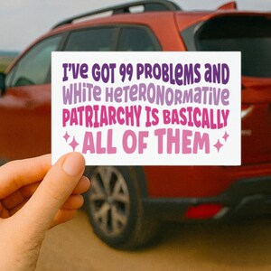May include: A white rectangular sticker with the text "I'VE GOT 99 PROBLEMS AND WHITE HETERONORMATIVE PATRIARCHY IS BASICALLY ALL OF THEM" in purple and pink lettering. The sticker is held in front of a red car.