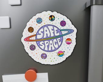 May include: A circular sticker featuring a central purple planet with a ring and the text SAFE SPACE in white bubble letters. The design is surrounded by smaller planets with various patterns and colours, including stripes, swirls, and pride flag colours, set against a white background with small colourful stars.