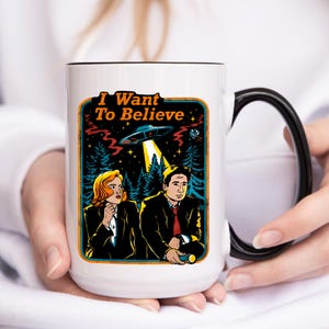 May include: White ceramic mug with a black handle and rim. The mug features a retro-style illustration with the text "I Want To Believe", a UFO, and two people in suits. The design is set against a forest backdrop.