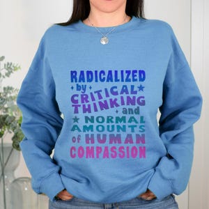 May include: A light blue crewneck sweatshirt featuring a multi-colored graphic print with the text: RADICALIZED by CRITICAL THINKING and NORMAL AMOUNTS of HUMAN COMPASSION. The lettering transitions from blue to purple, teal, and pink hues.
