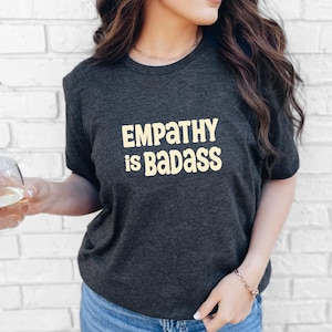 May include: Dark gray t-shirt with the text "EMPATHY is BADASS" in a light yellow font. The shirt has a crew neck and short sleeves. The background is a white brick wall.