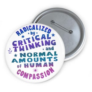 Può includere: Un bottone bianco con la scritta "RADICALIZED by CRITICAL THINKING and NORMAL AMOUNTS of HUMAN COMPASSION" in blu, viola e verde acqua. Il bottone ha un retro in metallo argentato e una spilla di sicurezza.