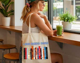 May include: A beige canvas tote bag featuring the text I READ BANNED BOOKS in blue letters above a graphic illustration of a row of colorful book spines with various titles including The Handmaid's Tale, The Kite Runner, and To Kill a Mockingbird.