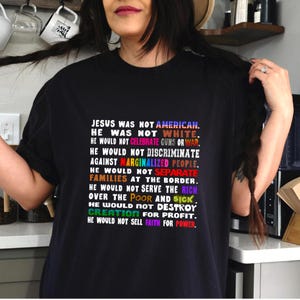 May include: A black t-shirt featuring multi-colored text that reads: JESUS WAS NOT AMERICAN. HE WAS NOT WHITE. HE WOULD NOT CELEBRATE GUNS OR WAR. HE WOULD NOT DISCRIMINATE AGAINST MARGINALIZED PEOPLE. HE WOULD NOT SEPARATE FAMILIES AT THE BORDER.