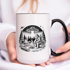 May include: White ceramic mug with a black handle. The mug features a black and white illustration of a Bigfoot and an alien sitting by a campfire in the woods. A UFO is flying overhead.