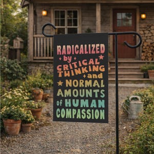 以下が含まれることがあります： 「RADICALIZED by CRITICAL THINKING and NORMAL AMOUNTS of HUMAN COMPASSION」というテキストがカラフルなグラデーション文字で書かれた黒いガーデンフラッグ。黒い金属製のスタンドに飾られており、背景には家と庭があります。